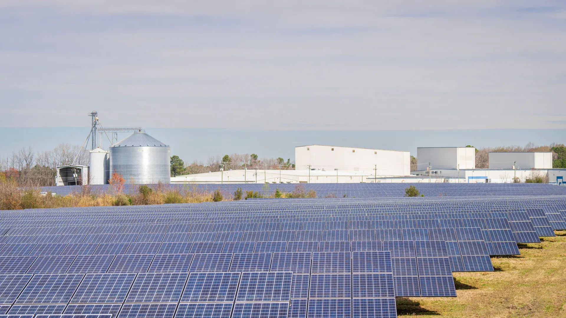 A large solar panel field with warehouses and silos in the background under a clear sky