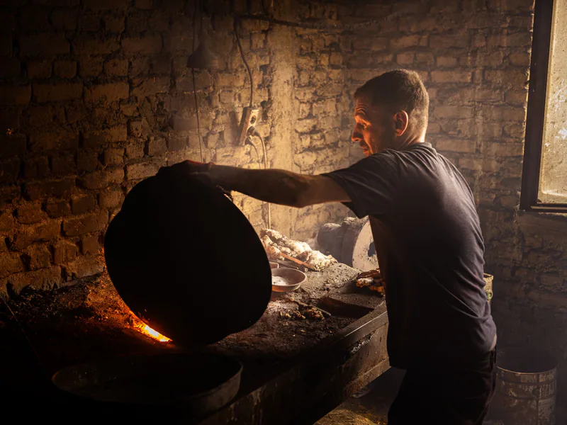 A craftsman works on copperware in a rustic indoor workshop, illuminated by warm firelight