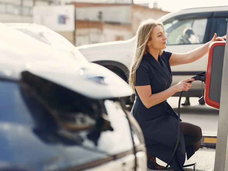 Side view of full length female driver in casual dress squatting near station while charging electric automobile for continuation of driving