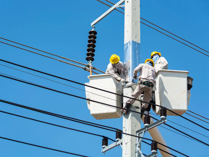 Electricians wearing safety gear work on power lines using lifts against a clear blue sky