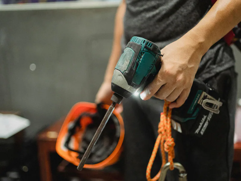 Crop unrecognizable mechanic wearing safety belt standing with switched on professional screwdriver and orange helmet before working indoors