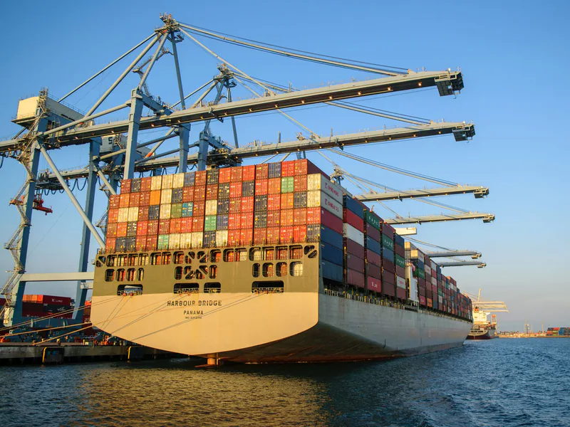 A large container ship at an industrial harbor with cranes under a clear blue sky